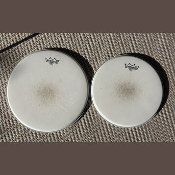 Dvě bílé bubnové kožehy značky REMO. Obě mají v horní části logo s nápisem REMO a pod ním text EMPEROR COATED MADE IN U.S.A. Povrchy kožehů vykazují…
