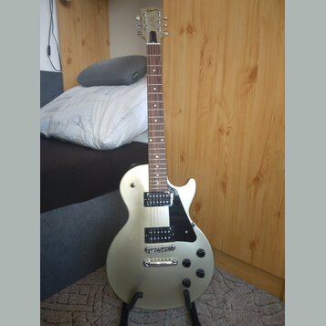 Stříbrná elektrická kytara s černým pickguardem a dvěma humbuckery. Na hlavě hmatníku je viditelné logo Gibson. Nástroj je umístěn ve stojanu v inter…