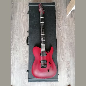 Červená elektrická kytara s matným povrchem položená na černém tvrdém kufru. Na hlavě hmatníku je bílý nápis "Schecter". Nástroj má dva humbuckery, j…