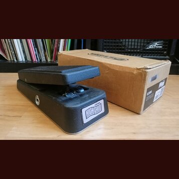 Černý volume pedál s nápisem VOLUME HIGH GAIN na bílém štítku. Vedle pedálu je umístěna hnědá kartonová krabice s nápisem GCB80 a čárovým kódem. Povr…