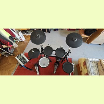 Elektronická bicí souprava s černými pady a činely na černém kovovém rámu. Na modulu je nápis Roland, stejně jako na bílém rámech basového bubnu. Sou…