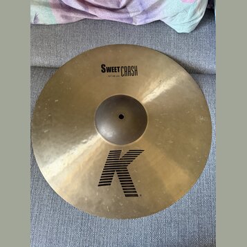 Zlatavý mosazný činel s viditelným kovánívým povrchem. Na horní části je černý nápis SWEET CRASH a pod ním menší text 19"/48 cm. Ve spodní části je v…