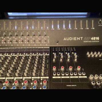 Fotografie zachycuje část míchacího pultu s nápisem AUDIENT ASP 4816 HERITAGE EDITION. Zařízení má šedý kovový povrch a je vybaveno řadou posuvníků,…