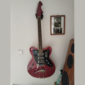 Červená elektrická kytara s polodupatým korpusem a f-otvory, zavěšená na bílé stěně. Nástroj má tmavý hmatník, černý pickguard a stříbrný vibrační sy…
