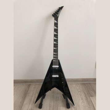 Černá elektrická kytara s tělem ve tvaru V umístěná na stojanu. Na hlavě hmatníku je bílý nápis Jackson. Kytara má dva humbuckery, jeden ovládací kno…