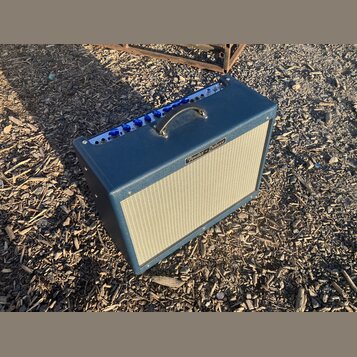 Kytarový kombozesilovač s tmavě modrým potahem a světlou textilní maskou reproduktoru. Na předním panelu je nápis "Fender Deluxe" a horní ovládací pa…