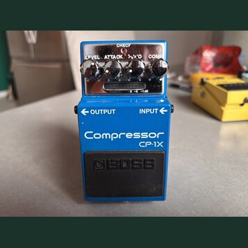 Modrý efekt pedál s nápisy Compressor a CP-1X. Na horní části jsou čtyři ovládací knoflíky s popisky LEVEL, ATTACK, 'V'O a COMP, kontrolka CHECK a př…