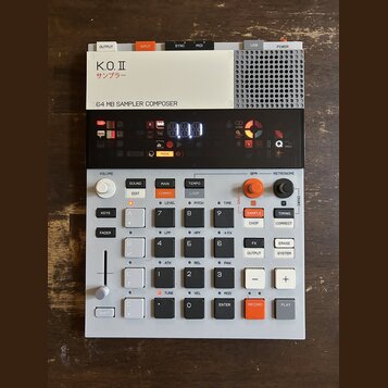 Bílo-šedý sampler s černým displejem a oranžovými detaily. Na horním bílém panelu je nápis K.O.II, pod ním japonské znaky サンプラー a text 64 MB SAMPLER…