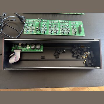 Fotografie zobrazuje dvě zelené elektronické desky s osazenými součástkami a černý obdélníkový šasi z tmavého dřeva. Na jedné z desek je vidět nápis…