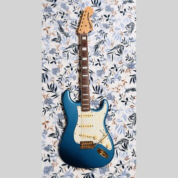 Modrá elektrická kytara s bílým perleťovým pickguardem a zlatými hardwaremi. Na hlavě hmatníku je nápis Squier STRATOCASTER. Hmatník je tmavého dřeva…
