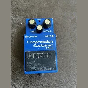 Modrý efekt pedál s nápisy LEVEL, SUSTAIN, OUTPUT a INPUT. Na přední straně je bílý text Compression Sustainer CS-2 a černé logo BOSS. Zařízení vykaz…