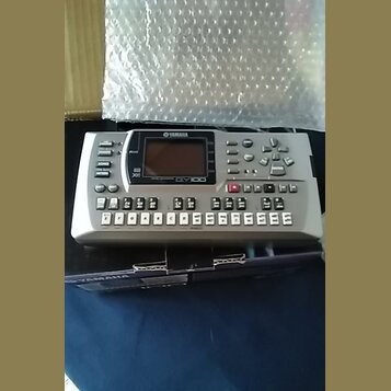 Stříbrný elektronický sekvencer s displejem a mnoha ovládacími tlačítky. Na přístroji jsou nápisy YAMAHA, QY100 a XBass. Zařízení leží na tmavém podk…