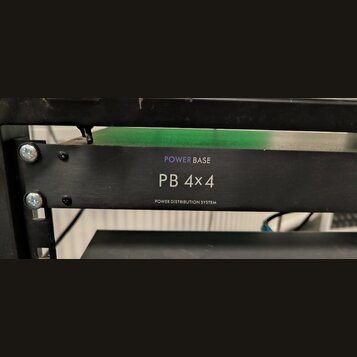 Černý rackový rozvodč rač s nápisy POWER BASE, PB 4x4 a POWER DISTRIBUTION SYSTEM. Zařízení je přišroubované do rackového stojanu pomocí čtyř šroubů.…