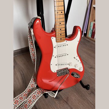 Červená elektrická kytara typu Stratocaster s bílým pickguardem a světle hnědým javorovým hmatníkem. Nástroj má tři snímače, tři ovládací knofíky a c…