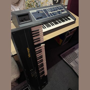 Na fotografii jsou dva klávesové syntezátory. V horní části je šedý přístroj s nápisy Emulator II a E-mu Systems, inc., který má v horní části ovláda…