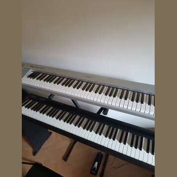 Dvě klávesové nástroje umístěné nad sebou na černých stojanech. Horní instrument je stříbrný s nápisem YAMAHA na horním panelu. Dolní instrument je č…