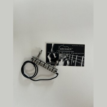 Kovový tremolo systém pro kytaru s černým přívodním kabelem. Vedle nástavce leží vizitka s nápisy BRENNER™, WWW.BRENNERUSA.COM, PRECISION GUITAR PROD…