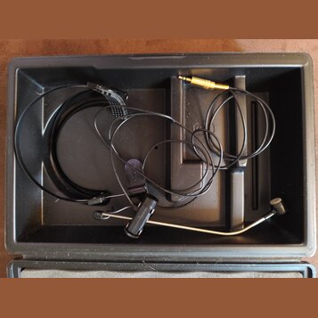 Černý plastový kufřík s vnitřními přihrádkami, ve kterém se nachází headset s mikrofonem. Sestava obsahuje tenký černý kabel, mikrofonní rameno s čer…