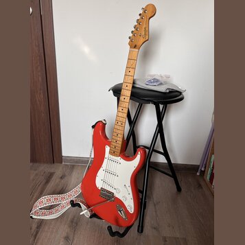 Červená elektrická kytara typu Stratocaster s bílým pickguardem a světle hnědým hmatníkem. Na hlavě kytary je nápis Squier by Fender STRATOCASTER. Ná…