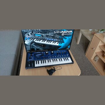 Modrý kompaktní syntetizátor s bílými klávesami a černými ovládacími prvky. Na přístroji je nápis MININOVA 2 a logo novation. Syntetizátor stojí před…
