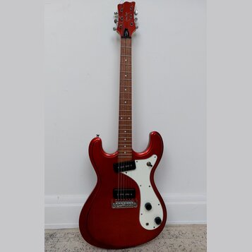 Elektrická kytara s červeným metalickým povrchem těla a hnědým hmatníkem. Na hlavě je bílý nápis "Cort". Kytara má bílý pickguard, jeden snímač typu…