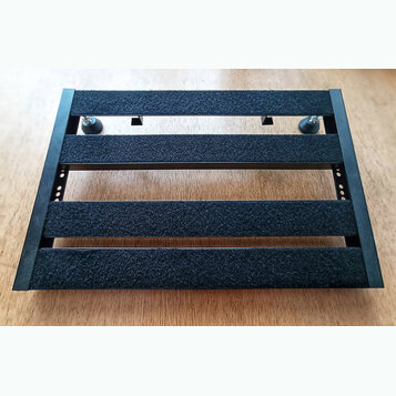 Černý obdélníkový pedalboard s třemi vodorovnými lištami potaženými černým hrubým povrchem. Boční rám je vyroben z černého materiálu s viditelnými ot…