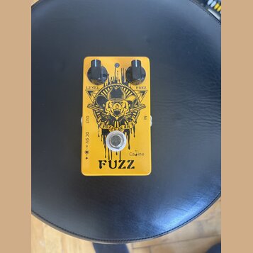 Žlutý efekt pedál s nápisy LEVEL a FUZZ u dvou černých ovládacích knoflíků. Uprostřed je grafika medvěda v klobuku s nápisem FUZZY BEAR a v dolní čás…