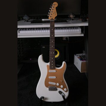 Bílá elektrická kytara typu Stratocaster s hnědým pickguardem a hnedým hmatníkem. Na hlavě je vidět nápis Fender. Kytara má tři snímače, tři ovládací…