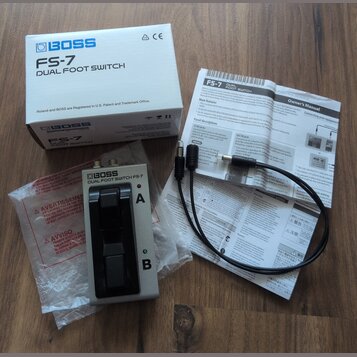 Na fotografii je stříbrný pedál BOSS DUAL FOOT SWITCH FS-7 s dvěma černými spínači. K pedálu patří původní bílá krabice s nápisy BOSS FS-7 DUAL FOOT…