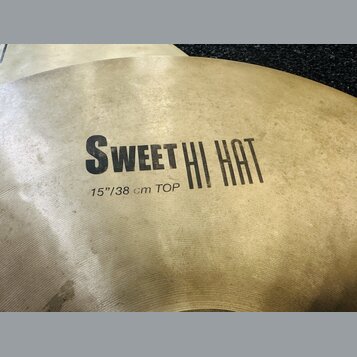 Detailní pohled na mosazný činel s viditelným broušením povrchu. Na čineli je černý nápis SWEET HI HAT a pod ním text 15"/38 cm TOP. Povrch vykazuje…