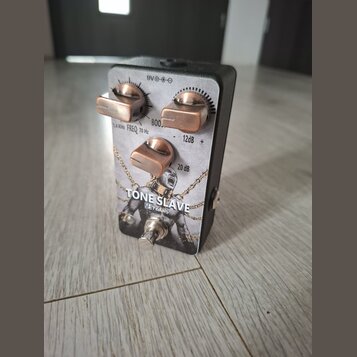 Efektový pedál s černým šasi a stříbrno-šedou přední deskou. Na desce je nápis TONE SLAVE a logo VB Pedals, doplněné motivem postavy v řetězech. Pedá…
