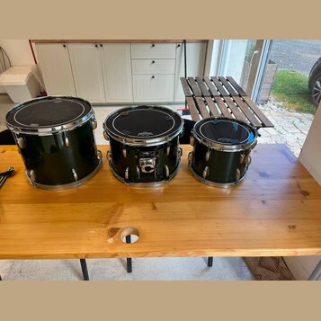 Tři černé bubny s kovovými obručemi a ladidly umístěné na dřevěném stole. Na každém z bubnů je v horní části bílý nápis "Gretsch". Povrch nástrojů je…