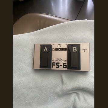 Stříbrný dvoupedálový spínač s černými plasty. Na horní straně je logo BOSS a nápisy DUAL FOOT SWITCH oraz FS-6. Pedály jsou označeny písmeny A a B,…