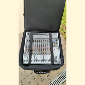 Stříbrný mixážní pult umístěný v černém polstrovaném přenosném pouzdře. Na zařízení jsou viditelné nápisy "MAIN MIX", "SUBGROUP" a řada číselných ozn…