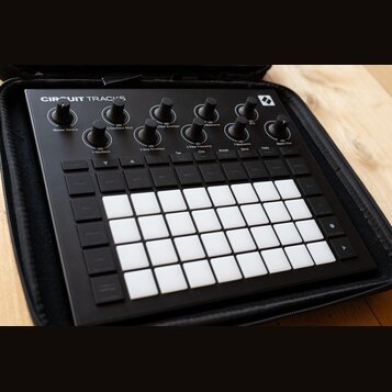 Černý sekvenční synthesizer Novation Circuit Tracks v černém přenosném pouzdře. Horní část obsahuje několik otočných ovladačů s bílými popisky jako M…