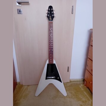 Bílá elektrická kytara s tělem ve tvaru V a hnědým hmatníkem. Na hlavě je viditelné zlaté logo Gibson. Tělo má černý plech s dvěma snímači a ovládací…