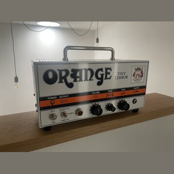 Kytarový zesilovač značky Orange, model Tiny Terror, v bílém provedení s kovovou rukojetí. Na předním panelu je velký černý nápis ORANGE a menší text…