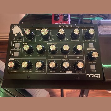 Černý analogový basový syntetizátor s nápisem MINITAUR a analog bass synthesizer. V levém dolním rohu je bílé logo moog. Přístroj obsahuje několik řa…