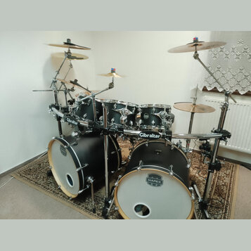 Černá bicí souprava s bílými لديmi a chromovaným kováním. Na basových bubnech je logo MAPEX, na tomarech jsou vidět loga Mapex a na nosném systému ná…