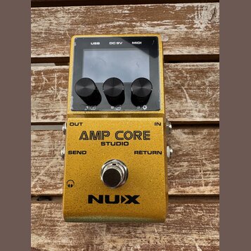 Zlatý efekt pedál s nápisy AMP CORE STUDIO a NUX. Horní část obsahuje displej, tři černé ovládací knoflíky a konektory USB, DC 9V a MIDI. Na spodní č…
