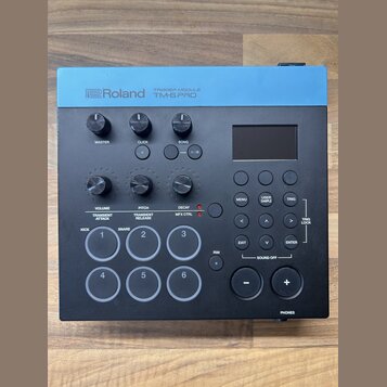 Černý trigger modul s modrým horním panelem. Na modré části je bílý nápis Roland TRIGGER MODULE TM-6 PRO. Zařízení obsahuje displej, několik otočných…