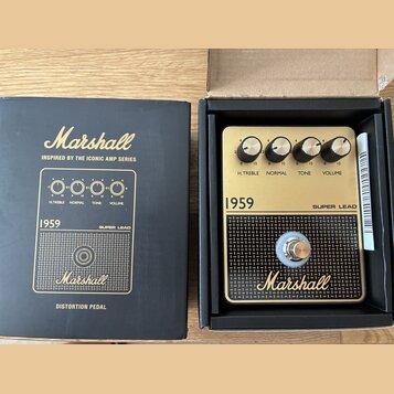 Černá krabice a v ní umístěný zlatý distortion pedál značky Marshall. Na pedálu jsou čtyři černé otočné ovladače s nápisy H.TREBLE, NORMAL, TONE a VO…