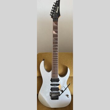 Elektrická kytara s bílým třpytivým povrchem těla a hnědým hmatníkem. Na hlavě je černý nápis Ibanez. Nástroj má tři humbuckery, tremolo mechanismus…