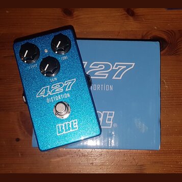 Modrý efekt pedál s glitrovým povrchem a tři ovládací knoflíky. Na pedálu jsou bílé nápisy 427 DISTORTION, LEVEL, GAIN, TONE a logo BBE. Vedle pedálu…