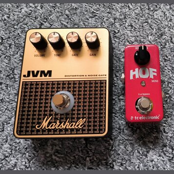 Dvě efektové pedály umístěné na šedém koberci. Levý pedál je zlatý s černým rámem, nápisy JVM, DISTORTION & NOISE GATE a Marshall, má čtyři ovládací…