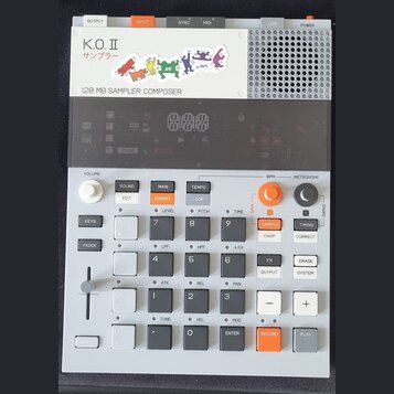 Šedý sampler s názvy K.O.II a nápisem サンプラー, pod kterým je text 128 MB SAMPLER COMPOSER. Zařízení obsahuje digitální displej, několik tlačítek v bílé…