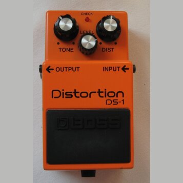 Oranžový efekt pedál s nápisy Distortion a DS-1. Na horní části jsou tři ovládací knoflíky s popisky TONE, LEVEL a DIST a červená kontrolka CHECK. Ve…