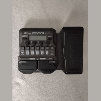 Černý multi-efektový procesor s nápisy A1X FOUR, ZOOM a Multi-Effects Processor. Zařízení obsahuje displej, několik ovládacích tlačítek s popisky jak…