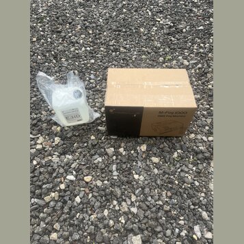Kartonová krabice s nápisy STARVLUX, M-Fog 1000 a DMX Fog Machine. Vedle krabice je plastová láhev s bílou tekutinou v průhledném sáčku, na které je…