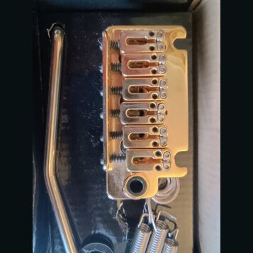 Zlatý Tremolo bridge pro elektrickou kytaru s šestnácti sedly a součástí je páka. Kovový povrch vykazuje známky opotřebení, drobné škrábance a oxidac…